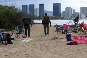 Officers from the Department of Land and Natural Resources Division of Conservation and Resource Enforcement agency walk along the beach of Sand Island State Recreation Area amid belongings of homeless camps.