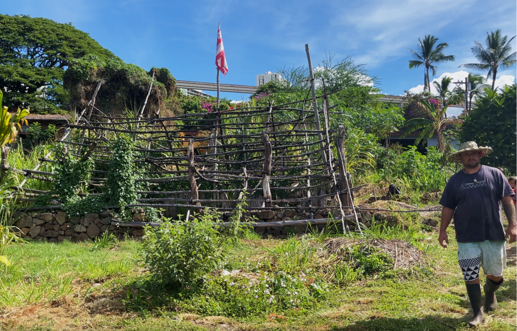 the wooden framework of a hale stands with the hae Hawaiʻi flying atop while a farmer strides forward in a sun hat and boots