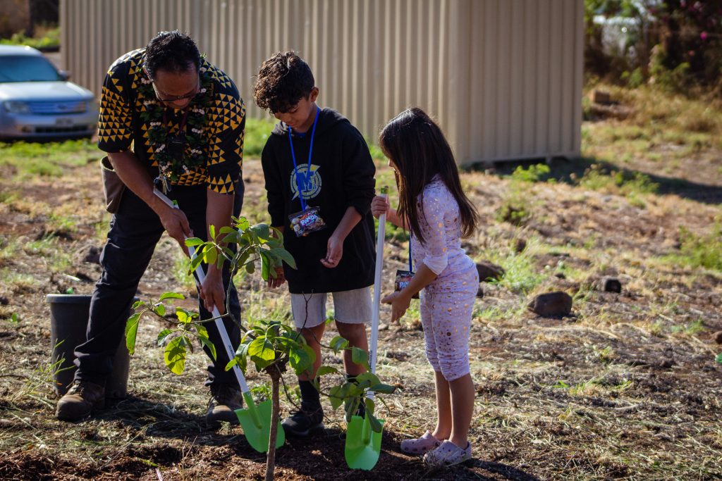 middle aged man helps two keiki shovel next to a newly planted tree
