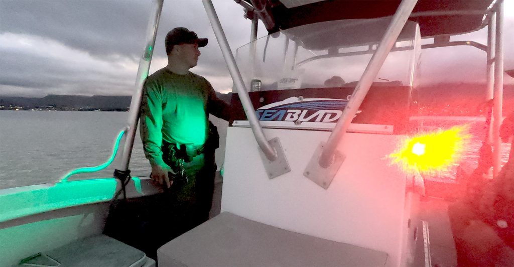 Picture taken from the bow of a DOCARE marine patrol boat on Kāne‘ohe Bay sandbar at dusk. DOCARE officer stands near the helm with the boat's bright green and red lights on and the bay waters and overcast sky in the background, 01-07-2022.