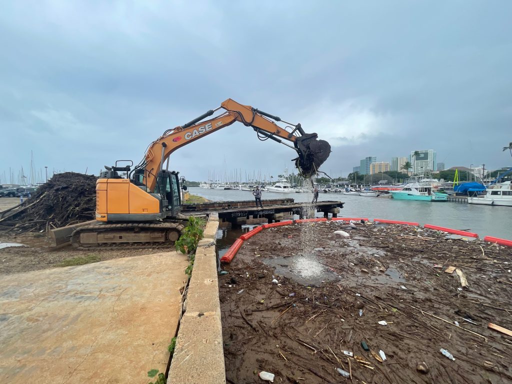 An excavator on the bank of the Ala Wai Canal lifts tree branches and sticks from the water where they are contained by a boom used to create a debris trap to prevent trash from entering the ocean, 03-15-2026.