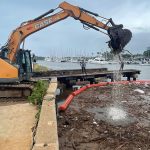 An excavator on the bank of the Ala Wai Canal lifts tree branches and sticks from the water where they are contained by a boom used to create a debris trap to prevent trash from entering the ocean, 03-15-2026.