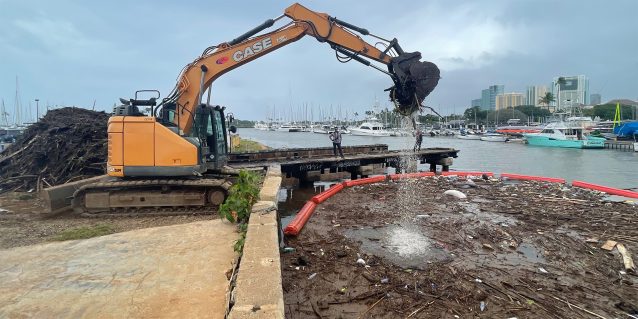 An excavator on the bank of the Ala Wai Canal lifts tree branches and sticks from the water where they are contained by a boom used to create a debris trap to prevent trash from entering the ocean, 03-15-2026.