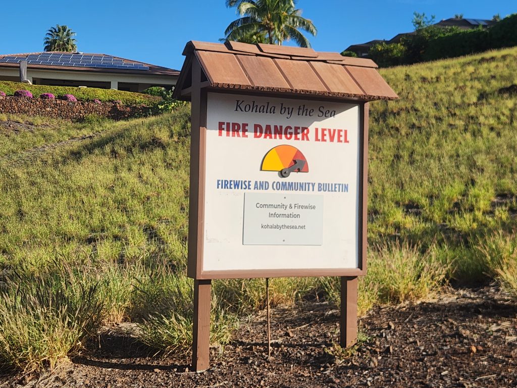 Wooden sign on hillside reading "Kohala by the Sea Fire Danger Level, Firewise and Community Bulletin." Graph with yellow, orange, and red colors with black arrow pointing to red.