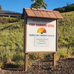 Wooden sign on hillside reading "Kohala by the Sea Fire Danger Level, Firewise and Community Bulletin." Graph with yellow, orange, and red colors with black arrow pointing to red.