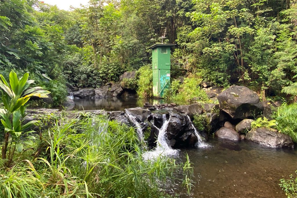 A two-story, narrow, green, rectangular structure which houses stream gauge equipment stands on the right side of Heʻeia Stream in Haiku Valley near Kāneʻohe, Oʻahu, just above a short drop-off over stream rocks.