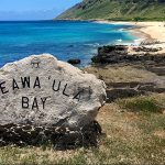 Photo of the rock with "Keawa‘ula Bay" carved on it, overlooking a crescent sandy beach on a sunny day at Ka’ena Point State Park, Aug-11-2017.