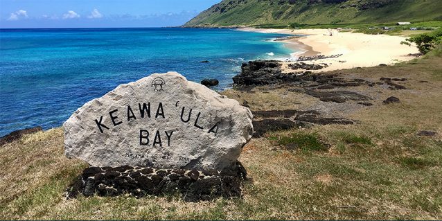 Photo of the rock with "Keawa‘ula Bay" carved on it, overlooking a crescent sandy beach on a sunny day at Ka’ena Point State Park, Aug-11-2017.