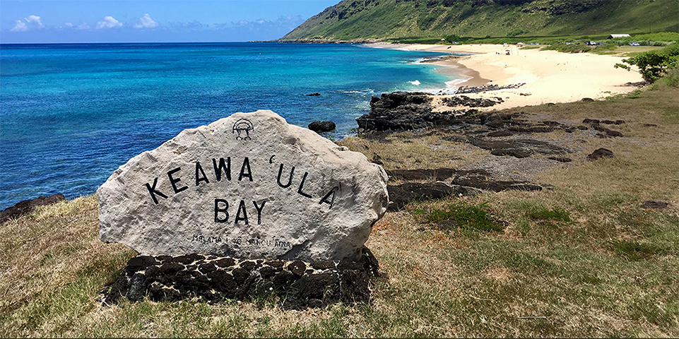 Photo of the rock with "Keawa‘ula Bay" carved on it, overlooking a crescent sandy beach on a sunny day at Ka’ena Point State Park, Aug-11-2017.