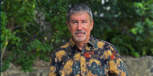Photo of Alan B. Carpenter standing in front of a garden with a low rock wall topped by tropical plants, July 27, 2026.